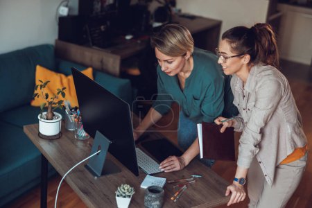 Foto de Trabajo en equipo, dos mujeres freelancer hablando mientras trabajan juntas en la computadora, haciendo proyecto en la oficina en casa. - Imagen libre de derechos