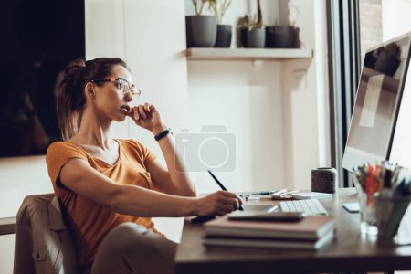 Téléchargez les photos : Jeune femme pigiste regardant réfléchi tout en travaillant sur l'ordinateur de son bureau à la maison. - en image libre de droit