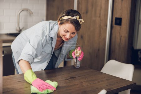 Photo for Mature woman smiling while cleaning a kitchen table at home. She is wearing protective gloves and using a spray bottle. - Royalty Free Image
