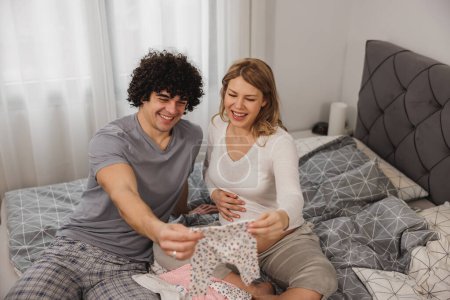 Téléchargez les photos : Joyeux couple enceinte regardant des vêtements de bébé tout en se relaxant sur un lit dans la chambre. - en image libre de droit