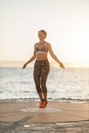 Foto de Fit middle age woman jumping rope during hard training near the sea beach. - Imagen libre de derechos