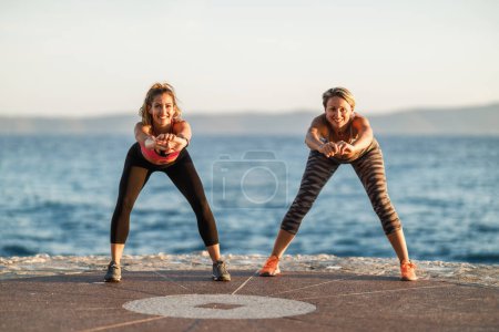 Photo for Two women friends are doing stretching exercises during training near the sea beach. - Royalty Free Image