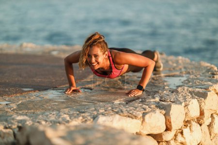 Foto de Mujer joven feliz haciendo ejercicios push-up durante el entrenamiento duro cerca de la playa del mar. - Imagen libre de derechos