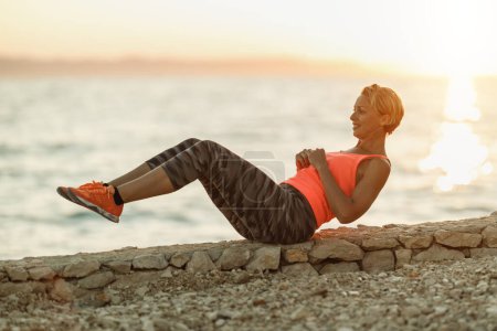 Foto de Ajuste mujer de mediana edad haciendo ejercicios de sentada durante el entrenamiento duro cerca de la playa de mar. - Imagen libre de derechos