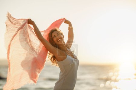 Foto de Una joven atractiva se divierte y disfruta de unas vacaciones de verano. Ella está posando con una bufanda transparente en sus manos en la playa. - Imagen libre de derechos