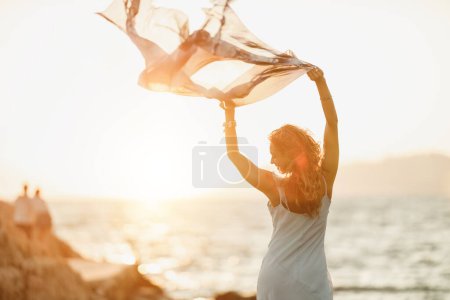 Photo for An attractive young woman is having fun and dancing with transparent scarf in her hands while enjoying a summer vacation on the beach. - Royalty Free Image