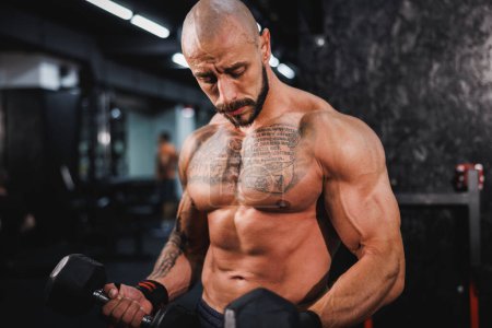 Téléchargez les photos : Shot of a muscular bodybuilder doing hard training with dumbbell at the gym. He is pumping up his biceps muscle with heavy weight. - en image libre de droit