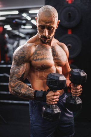 Foto de Shot of a muscular bodybuilder doing hard training with dumbbell at the gym. He is pumping up his biceps muscle with heavy weight. - Imagen libre de derechos