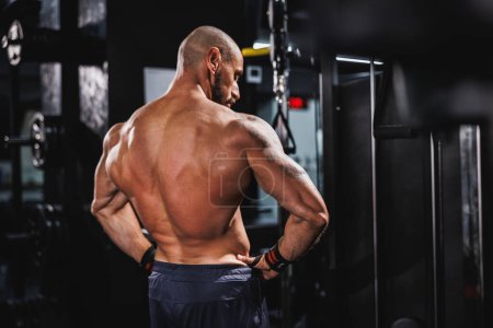 Téléchargez les photos : Vue arrière d'un jeune bodybuilder musculaire montrant ses muscles parfaits du dos tout en posant après avoir travaillé à la salle de gym. - en image libre de droit