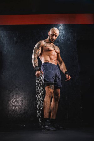 Téléchargez les photos : Un jeune homme de remise en forme debout dans la salle de gym et se préparant à la musculation avec une chaîne lourde. - en image libre de droit