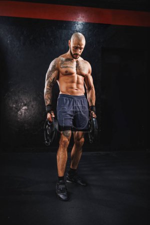 Téléchargez les photos : Prise de vue d'un homme musclé montrant ses muscles parfaits tout en se préparant à la musculation avec une plaque de poids à la salle de gym. - en image libre de droit
