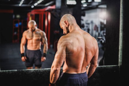 Photo for Rear view of a young fit Caucasian man who is posing in front of a mirror after working out in the gym. - Royalty Free Image