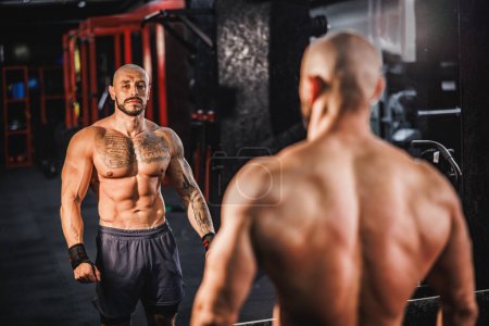 Téléchargez les photos : Prise de vue d'un jeune homme de fitness montrant son corps musclé parfait et six pack devant un miroir dans la salle de gym. - en image libre de droit