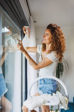 Téléchargez les photos : Femme souriante qui nettoie la fenêtre ou essuie le verre dans un appartement, une maison ou un hôtel. - en image libre de droit