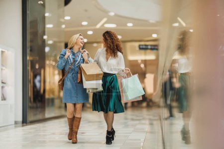 Foto de Fotografía de dos amigas alegres caminando por un centro comercial mientras llevan bolsas. - Imagen libre de derechos