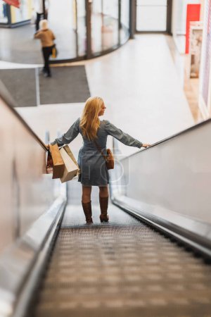 Téléchargez les photos : Vue arrière d'une belle femme sur l'escalator dans un centre commercial lors d'une virée shopping. - en image libre de droit