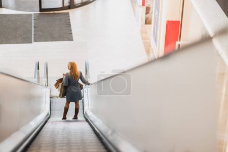 Téléchargez les photos : Vue arrière d'une jolie femme portant des sacs sur un escalier roulant dans un centre commercial lors d'une virée shopping. - en image libre de droit