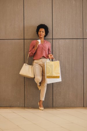 Téléchargez les photos : Prise de vue d'une femme noire portant des sacs à provisions et tenant une tasse de café à emporter debout contre un mur dans un centre commercial. - en image libre de droit