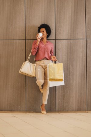 Photo for Shot of a Black woman drinking coffee to go while while holding shopping bags against a wall in a mall. - Royalty Free Image