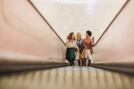 Foto de Tiro de tres mujeres de cheerfu divirtiéndose y de pie en escaleras mecánicas en un centro comercial de la ciudad juntos mientras que hacia fuera en una juerga de compras. - Imagen libre de derechos