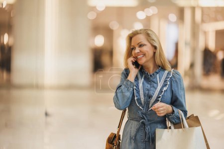 Téléchargez les photos : Femme souriante parlant sur son smartphone tout en marchant dans un centre commercial et en portant des sacs. - en image libre de droit