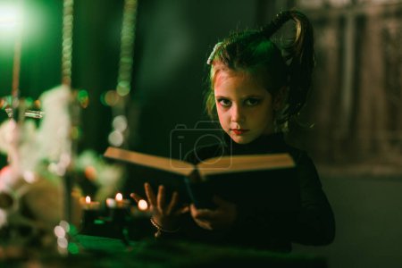 Téléchargez les photos : Portrait d'une jolie petite fille avec un énorme vieux livre qui se prépare pour Halloween. Regardant la caméra. - en image libre de droit