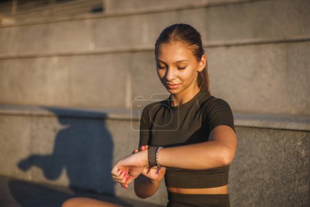 Foto de Una joven deportista que mira en smartwatch y descansa después de un duro entrenamiento al aire libre. - Imagen libre de derechos