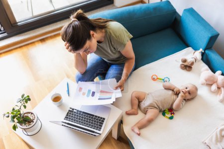 Téléchargez les photos : Tourné d'une jeune mère occupée regardant stressé tout en travaillant à la maison tout en prenant soin de son adorable bébé. - en image libre de droit