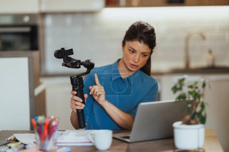 Téléchargez les photos : Femme vlogging sur les produits de photographie et montrant cardan pour appareil photo tout en ayant réunion en ligne sur son ordinateur portable à la maison. - en image libre de droit