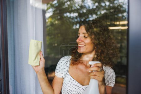 Téléchargez les photos : Femme souriante essuyant la vitre avec un chiffon dans un appartement, une maison ou un hôtel. Services d'entretien ménager ou d'accueil. - en image libre de droit