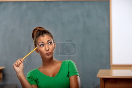 Photo for Portrait of a female student holding pencil and thinking while standing in front of a blackboard in the classroom with looks up. - Royalty Free Image