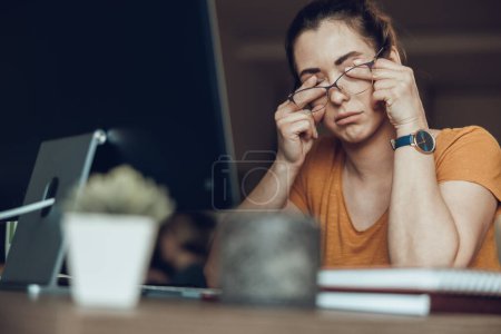 Photo for Sleepy young woman rubs her eyes while working on computer from her home office - Royalty Free Image