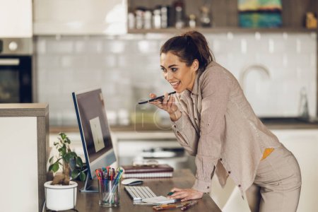 Téléchargez les photos : Jeune femme pigiste utilisant un smartphone tout en travaillant sur ordinateur à partir de son bureau à domicile. - en image libre de droit