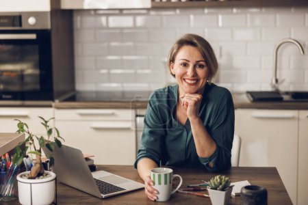 Téléchargez les photos : Portrait d'une femme mûre qui prend une pause café tout en travaillant à la maison. En regardant la caméra avec sourire. - en image libre de droit