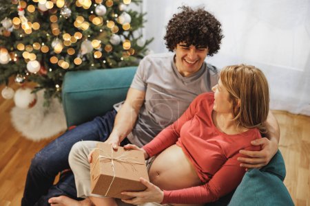 Foto de Joven pareja embarazada celebrando la Navidad en casa y dando regalos de vacaciones el uno al otro. - Imagen libre de derechos