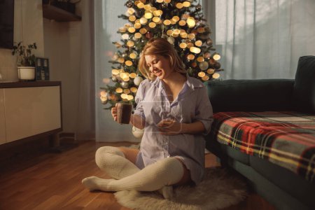 Foto de Mujer embarazada sentada en el suelo junto al árbol de Navidad y disfrutando de té o café. - Imagen libre de derechos