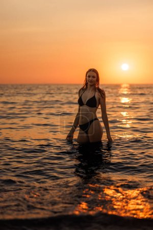 Foto de Hermosa mujer joven está de pie en la playa del mar en la puesta del sol. Ella agarra los últimos rayos del sol. - Imagen libre de derechos