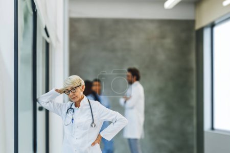 Photo for Tired senior female doctor looking worried while standing by the window while having a quick break in a hospital hallway. - Royalty Free Image
