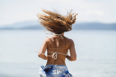 Téléchargez les photos : Vue arrière d'une jolie jeune femme en bikini s'amuse et danse sur la plage tout en profitant de vacances d'été. - en image libre de droit
