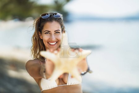 Téléchargez les photos : Belle jeune femme souriante tenant des étoiles de mer et profitant de la plage. Concentration sélective. Focus sur le fond, sur la fille. - en image libre de droit