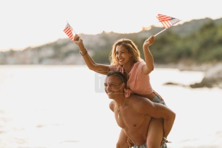 Téléchargez les photos : A loving cheerful couple with US national flag having fun while spending the day on the beach. - en image libre de droit