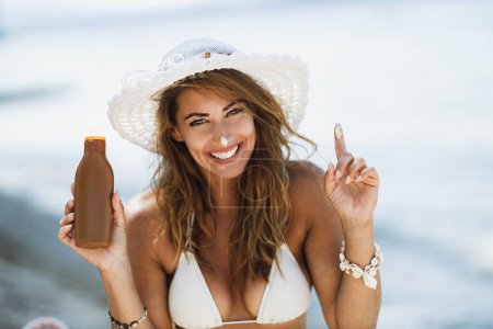 Foto de Una atractiva joven está sosteniendo crema de protección solar y disfrutando del tiempo en la playa. Mirando la cámara. - Imagen libre de derechos