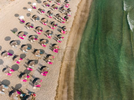 Luftaufnahme des erstaunlichen Sandstrandes mit Schilfschirmen und rosa Sonnenliege neben dem klaren Wasser des Mittelmeers an sonnigen Tagen.
