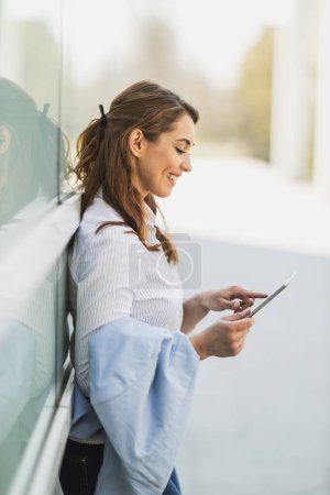 Foto de Joven mujer de negocios sonriente está navegando por la red social en la tableta digital en frente de la oficina durante el descanso. - Imagen libre de derechos
