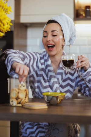 Téléchargez les photos : Jeune femme en peignoir manger des biscuits et boire du café le matin dans sa cuisine à la maison. - en image libre de droit