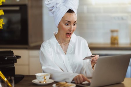 Téléchargez les photos : Une jolie femme en peignoir faisant du shopping en ligne sur son ordinateur portable tout en profitant du café du matin et se relaxant à la maison. - en image libre de droit
