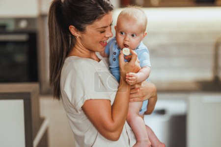 Téléchargez les photos : Jeune mère attentionnée qui s'amuse et nourrit son adorable bébé dans la cuisine à la maison. - en image libre de droit