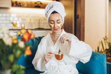 Foto de Una mujer atractiva con parches para el cuidado de la piel bajo los ojos disfrutando del té de la mañana y relajándose en su casa. - Imagen libre de derechos