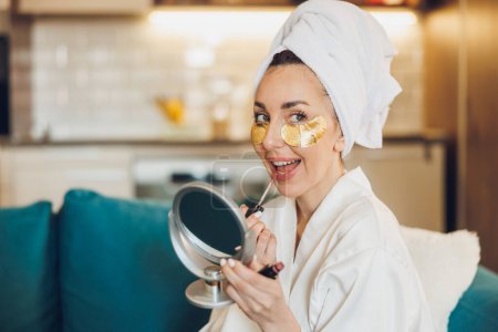 Foto de Una mujer atractiva con cuidado de la piel bajo parches para los ojos poniendo brillo labial mientras disfruta de la mañana en su casa. - Imagen libre de derechos