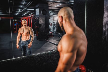 Téléchargez les photos : Prise de vue d'un jeune bodybuilder musculaire montrant ses muscles parfaits devant un miroir après avoir travaillé à la salle de gym. - en image libre de droit